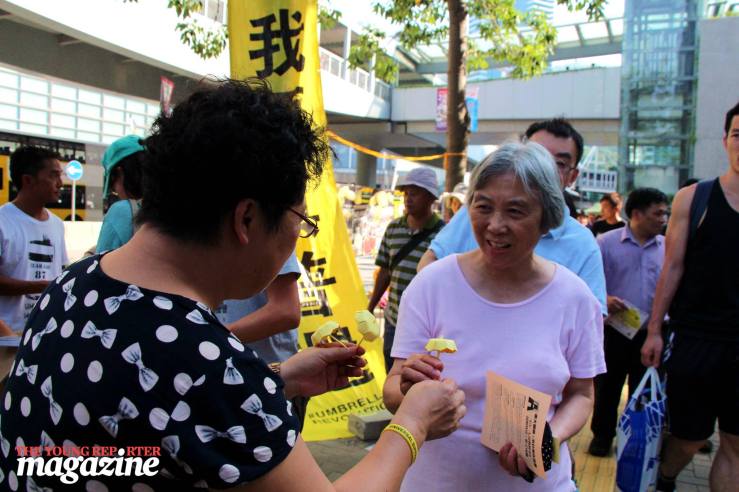 Woman hands out yellow umbrellas made of paper to passersby. | Legislative Council Complex | September 28, 2015 | Tanya McGovern